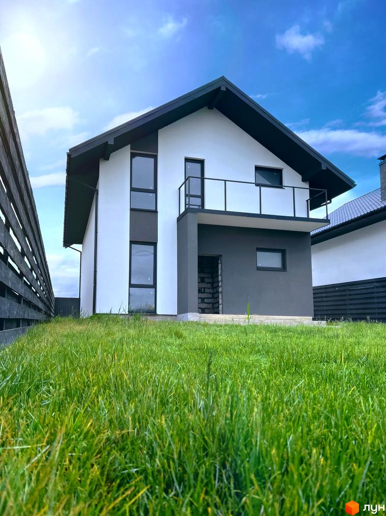 Котеджне містечко КМ Green Hill Village: двоповерховий будинок білого та сірого кольорів з балконом та темним дахом на тлі блакитного неба та зеленого газону.