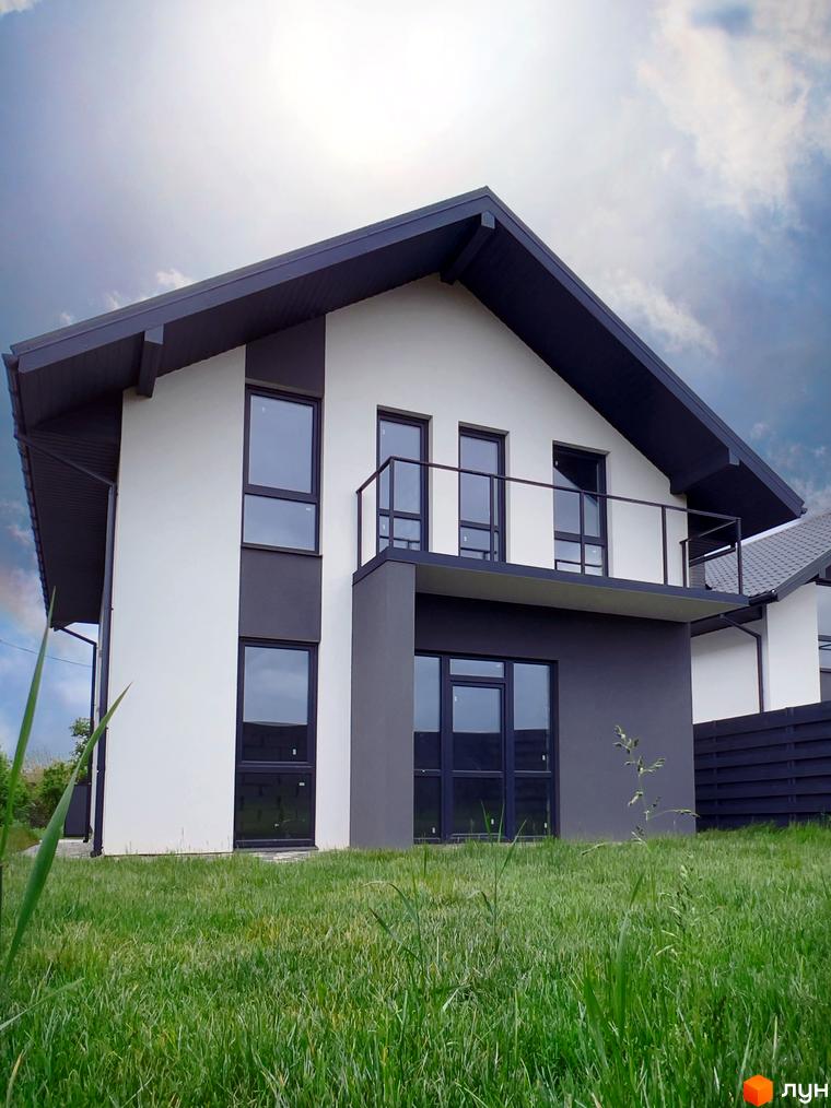 Двоповерховий будинок з темним дахом та вікнами, розташований на зеленій галявині. На балконі встановлені скляні перила, а на подвір'ї росте трава.