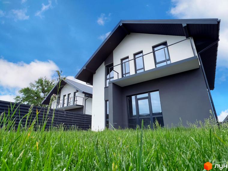Двоповерховий будинок з білими стінами та темним дахом у Містечку котеджів Green Hill Village. На передньому плані зелена трава, на задньому - блакитне небо з хмарами.
