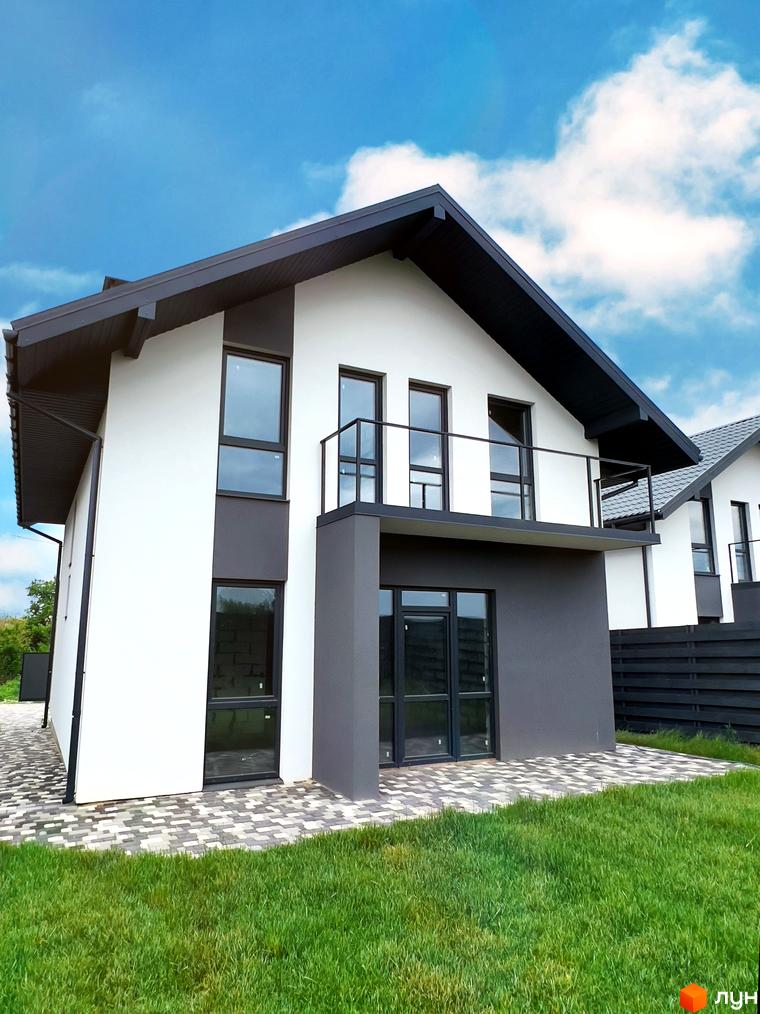Двоповерховий будинок з білими стінами та темним дахом. Перед будинком газон з травою та бруківка.