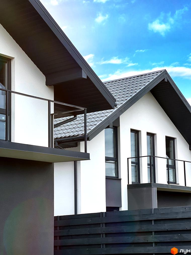 Котеджне містечко Green Hill Village: білі фасади будинків з темними дахами та вікнами, балкони зі скляними огорожами, дерев'яний паркан на фоні блакитного неба.