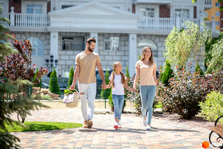 Сім'я гуляє біля Житлового комплексу Нова Англія. Фасад будинку з колонами та балконами, доглянута територія з бруківкою та зеленими насадженнями.