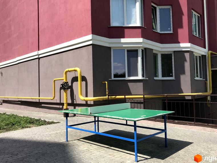 Житловий комплекс Святопетрівський: фасад будинку рожево-сірого кольору з білими вікнами. На прибудинковій території розташований зелений тенісний стіл на бруківці.