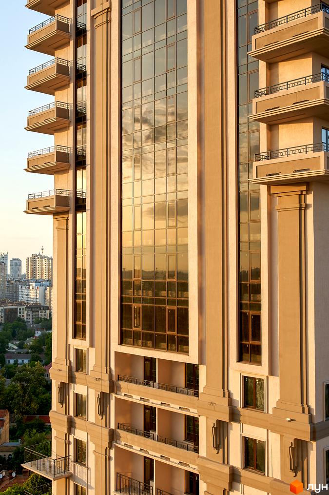 Фасад житлового комплексу Акрополь з великими скляними вікнами, що відбивають небо, та численними балконами. Навколо видно міські будівлі та дерева.