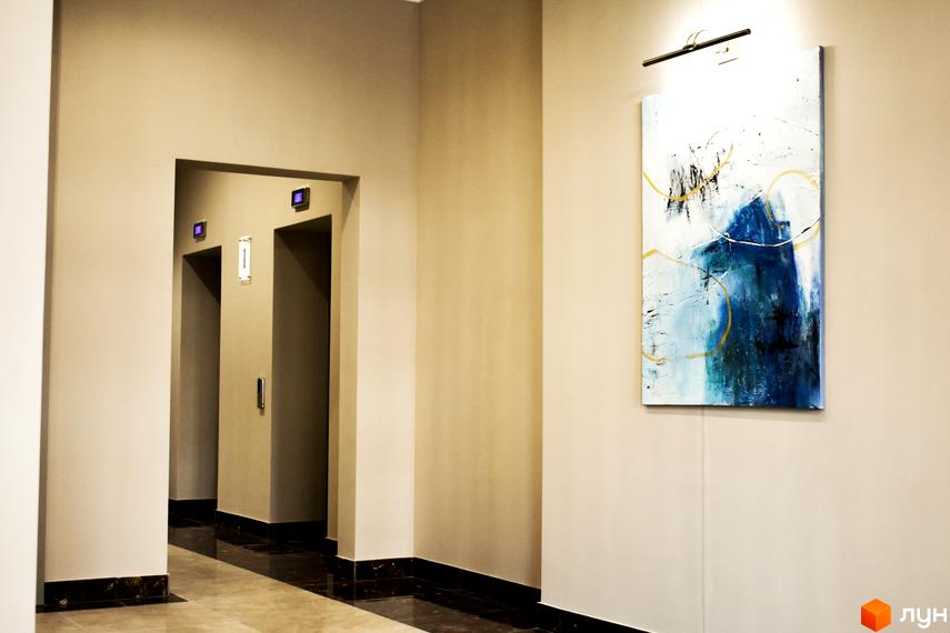 Інтер'єр холу в Житловому комплексі Акрополь з ліфтами та картиною на стіні. Коридор з кахлем на підлозі та чорним плінтусом.