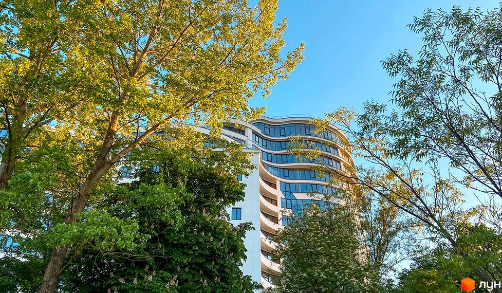 Зображення фасаду ЖК Parus на фоні зелених дерев та блакитного неба. Багатоповерховий будинок з хвилеподібними балконами.