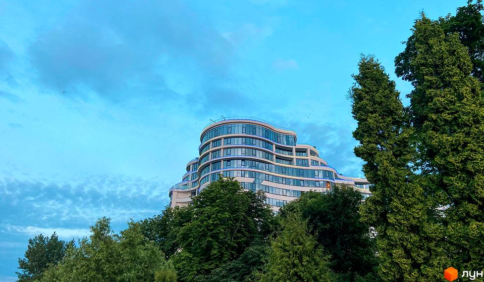 Фото фасаду Житлового комплексу Parus на фоні блакитного неба та зелених дерев. Вигляд багатоповерхового будинку з великими вікнами.