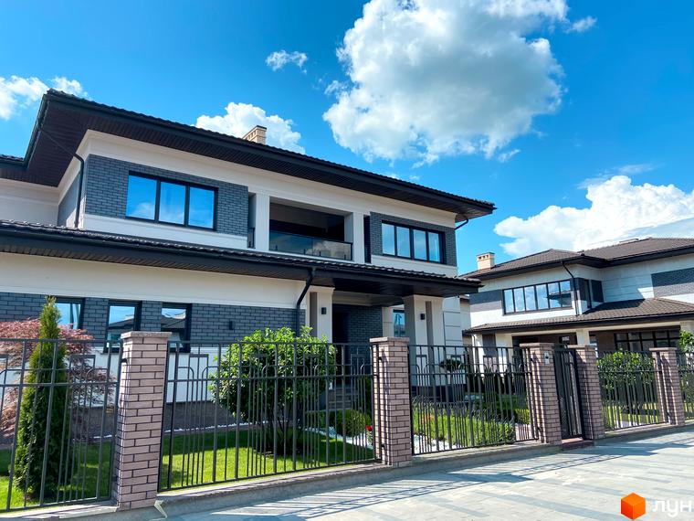 Котеджне містечко Franko River Club. Фасад будинку з сірої цегли та білої штукатурки, великі вікна, огороджена територія з зеленню та парканом.