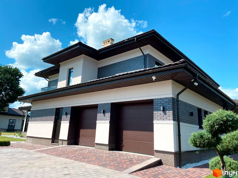 Котеджне містечко КМ Franko River Club з двома гаражними воротами та цегляним фасадом. Прибудинкова територія з доглянутим газоном та декоративним деревом.