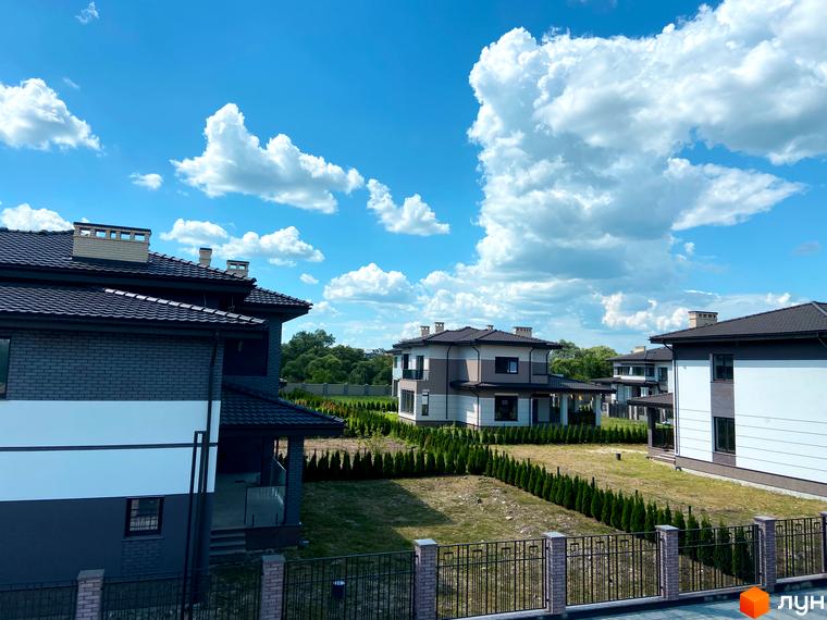 Котеджне містечко Franko River Club: фасади будинків з темними дахами та цегляними елементами на тлі блакитного неба з хмарами. Доглянута територія з туями та огорожею.