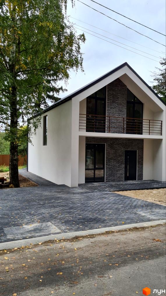 Зовнішній вигляд будинку в Котеджному містечку ForestHouse з балконом та сірою бруківкою на подвір'ї. Зелені насадження поруч.