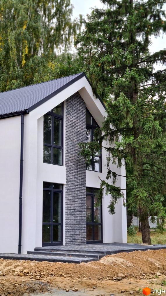 Котеджне містечко ForestHouse: Фасад будинку з білими стінами та сірою цегляною кладкою, великі темні вікна. Перед будинком вимощена територія та ґрунт.