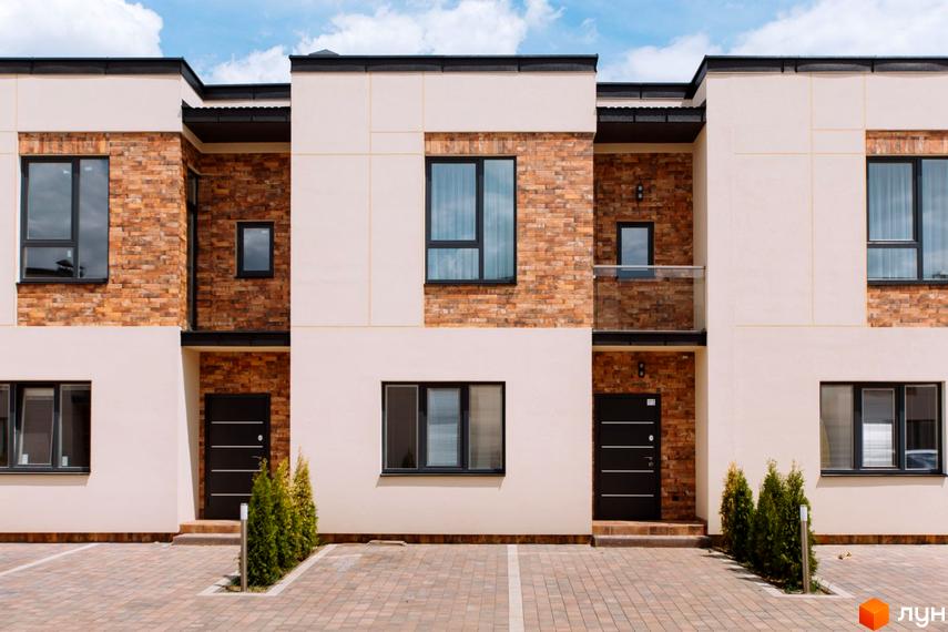 Фасад Котеджного містечка Малиновий з бежевими стінами та цегляними вставками, чорними вікнами і дверима. Перед будинками бруківка та зелені насадження.