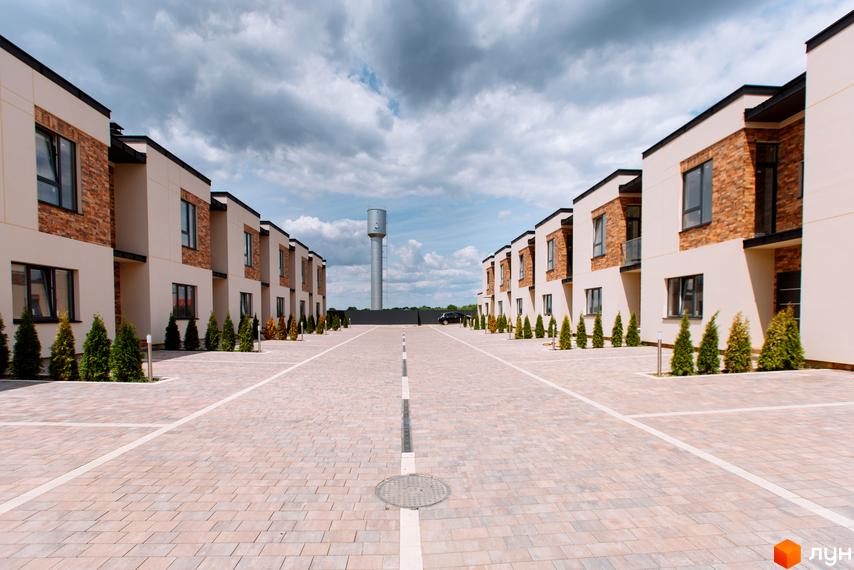 Ряд нових будинків з цегляним облицюванням та великими вікнами. Перед будинками розташована брукована площадка з зеленими насадженнями.