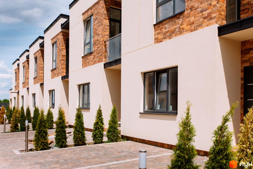 Зовнішній вигляд будинків Котеджне містечко Малиновий з білими стінами та коричневою цегляною обробкою. На прибудинковій території висаджені дерева та бруківка.