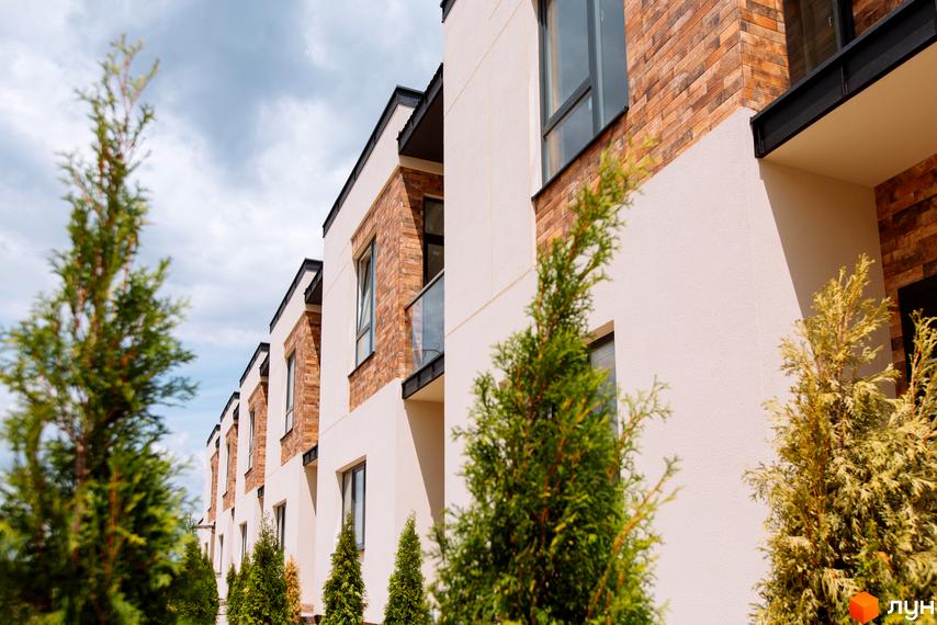 Котеджне містечко Малиновий: ряд котеджів з бежевими фасадами та цегляними вставками, перед якими висаджені зелені хвойні дерева.