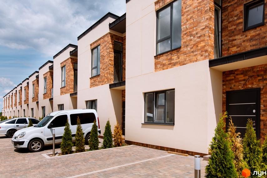 Ряд котеджів Котеджного містечка Малиновий з бежевими фасадами та цегляними вставками. Перед будинками облаштована парковка з автомобілями та декоративними деревами.