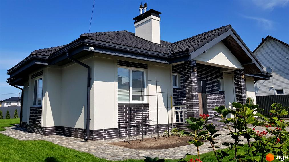 Котеджне містечко КМ Сонячний: двоповерховий будинок з темною черепицею, світлими стінами та цегляним оздобленням. Навколо будинку доглянутий газон та брукована доріжка.