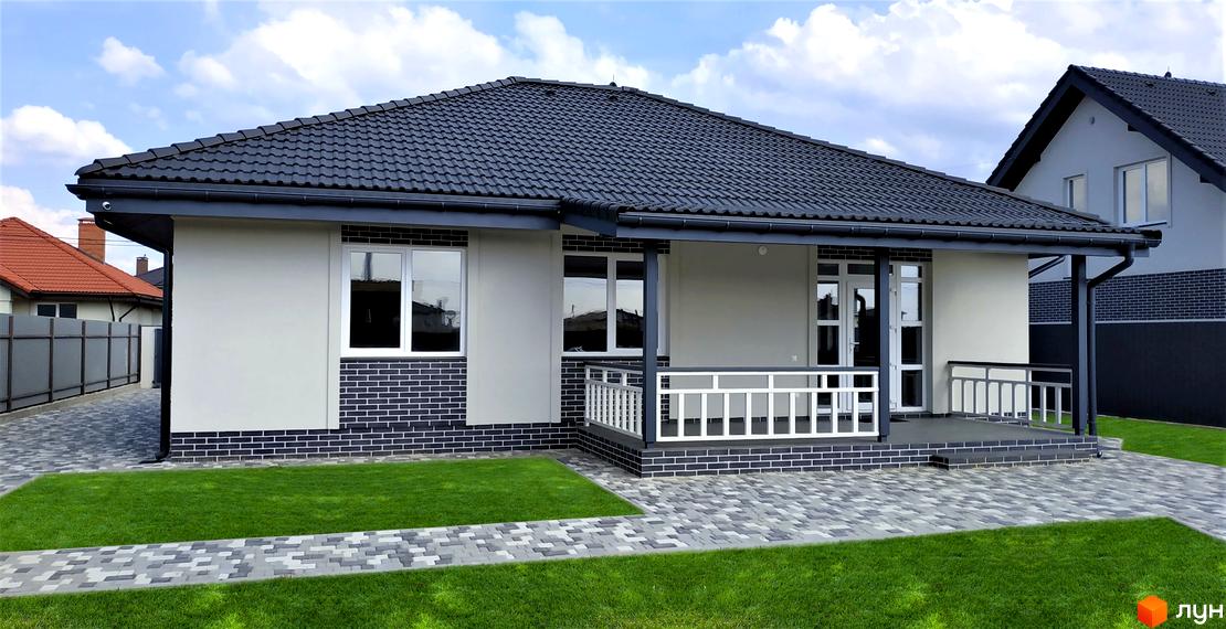 Котеджне містечко КМ Сонячний. Одноповерховий будинок з сірою черепичною покрівлею, світлими стінами та цегляним цоколем. Навколо будинку доглянутий газон та вимощені доріжки.