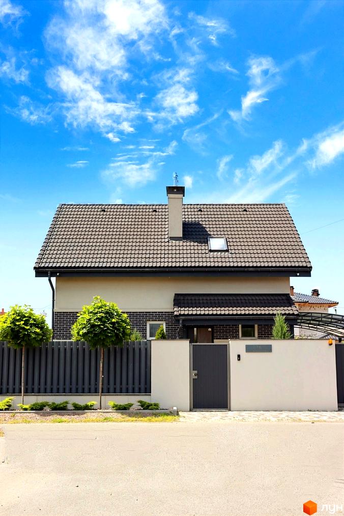 Котеджне містечко КМ Сонячний: двоповерховий будинок з коричневою черепицею та цегляною обробкою фасаду, огороджена територія з парканом та деревами.