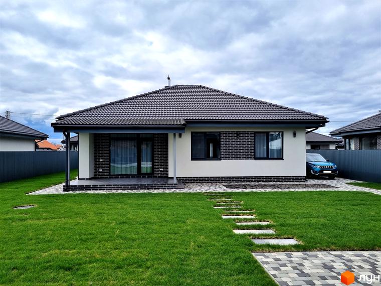 Котеджне містечко КМ Сонячний. Одноповерховий будинок з темним дахом, білими стінами та цегляними вставками. Зелений газон та доріжка з плитки перед будинком.