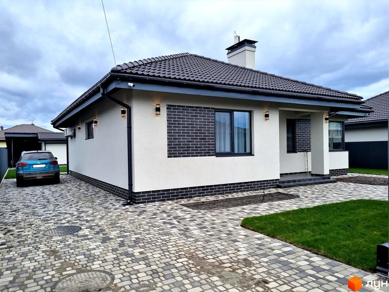 Котеджне містечко КМ Сонячний: одноповерховий будинок з білою штукатуркою, темною черепицею та цегляним декором. Прибудинкова територія з бруківкою та зеленим газоном.