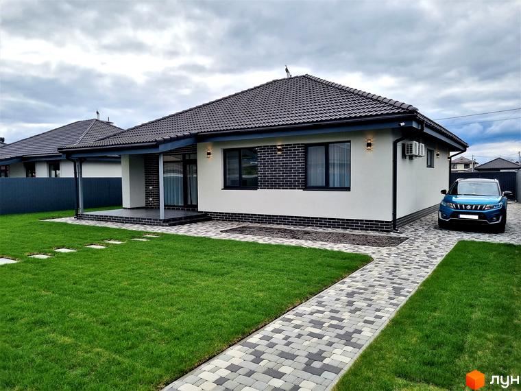 Котеджне містечко Сонячний. Одноповерховий будинок з темним дахом, білими стінами та цегляним оздобленням. Прибудинкова територія з доглянутим газоном та доріжкою з плитки.