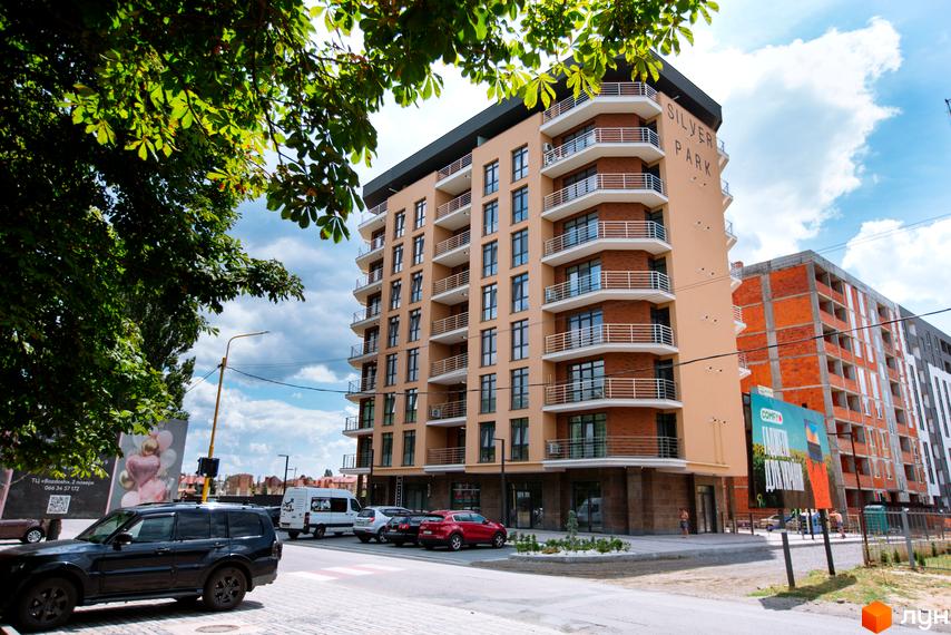 Фасад житлового комплексу SilverPark з балконами та вікнами, на тлі хмарного неба. Поруч розташовані автомобілі та дерева.