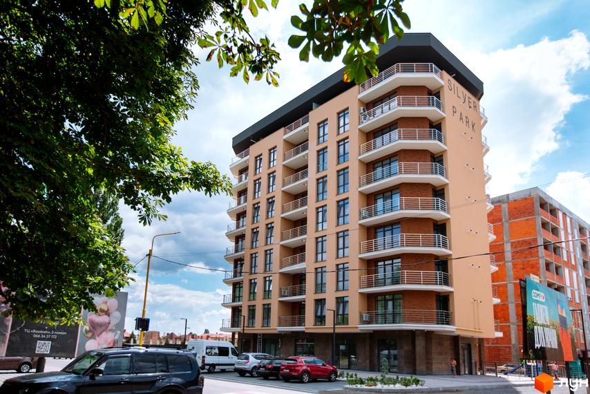 Житловий комплекс SilverPark – це багатоповерхова новобудова з балконами та панорамними вікнами. Поруч розташована прибудинкова територія з деревами та автомобілями.