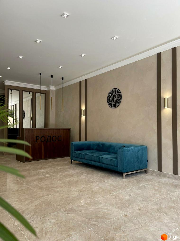 Вхідна група Житлового комплексу Родос з дерев'яною стійкою рецепції, бірюзовим диваном та світлою плитковою підлогою. На стіні круглий декор.