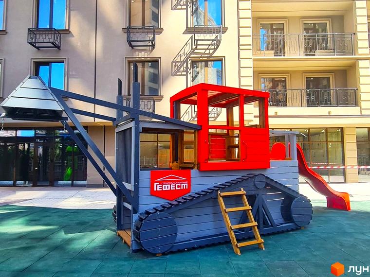 Фасад Житлового комплексу Родос світлого кольору з балконами та вікнами. На прибудинковій території розташований дитячий майданчик з ігровим комплексом у вигляді екскаватора.