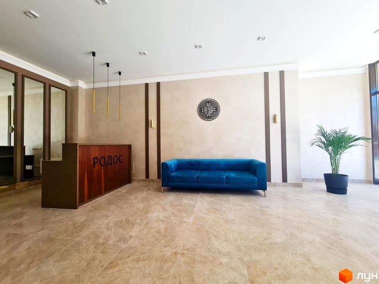 Лобі Житлового комплексу Родос з дерев'яною стійкою реєстрації та синім диваном. Інтер'єр доповнюють світлі стіни, плитка на підлозі та зелена рослина.