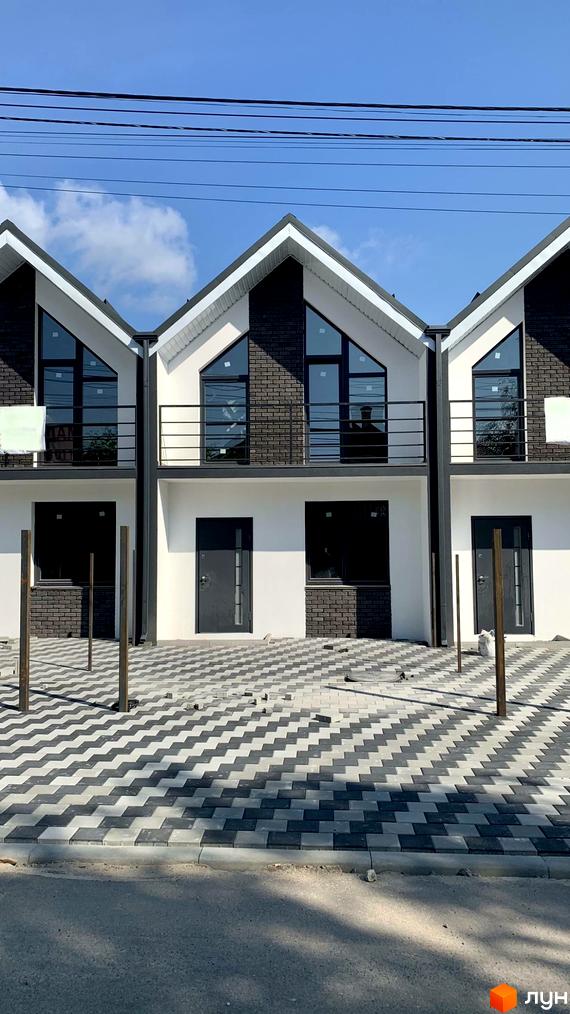 Триповерховий будинок з білими стінами та чорним дахом. Перед будинком викладена плитка у вигляді зигзагу, веде до асфальтної дороги.