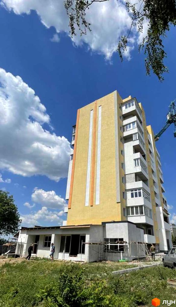 Висотна будівля ЖБК Добробут з жовтим фасадом та білими смугами. Поруч розташована прибудинкова територія з зеленню та будівлями на етапі будівництва.