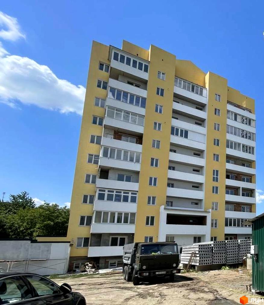 Висотна будівля ЖБК Добробут з жовтими фасадами та білими балконами. Поруч з будинком стоїть вантажівка та лежать будівельні матеріали.