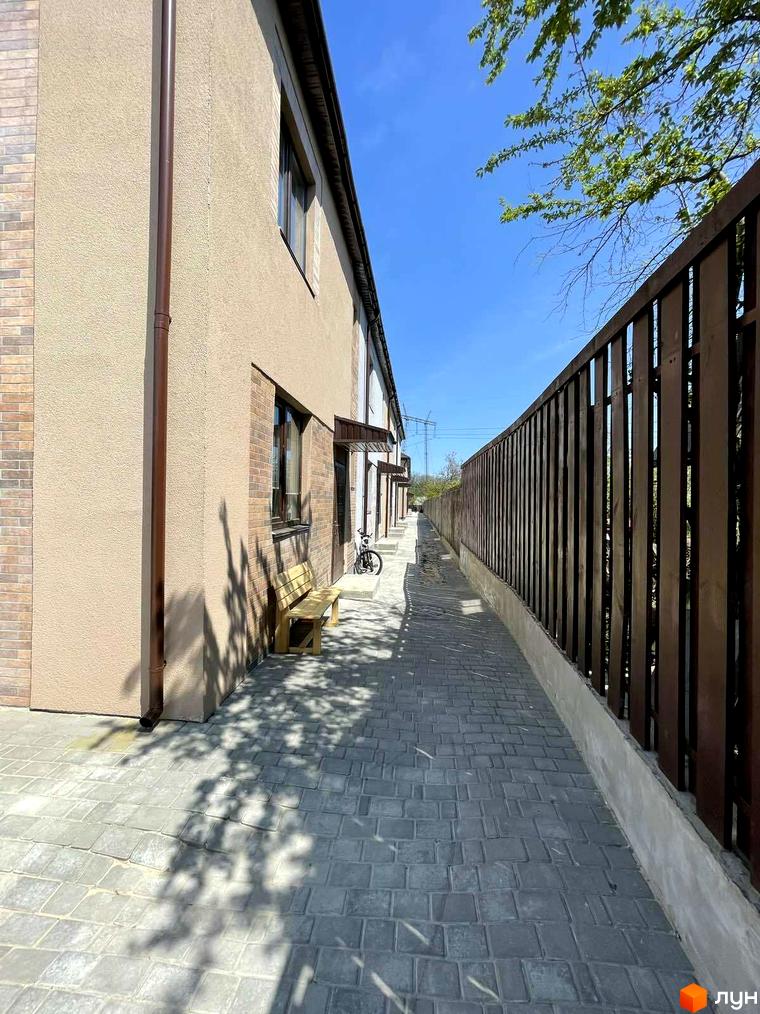 Вигляд фасаду таунхаусу Comfort home з коричневим водостоком та бруківкою. Поряд розташована вулична лава та огорожа з дерев'яних рейок.