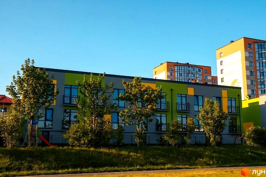 Котеджне містечко Дем’янів лаз з яскравим фасадом та зеленими деревами на прибудинковій території. На задньому плані видно багатоповерхові будинки.