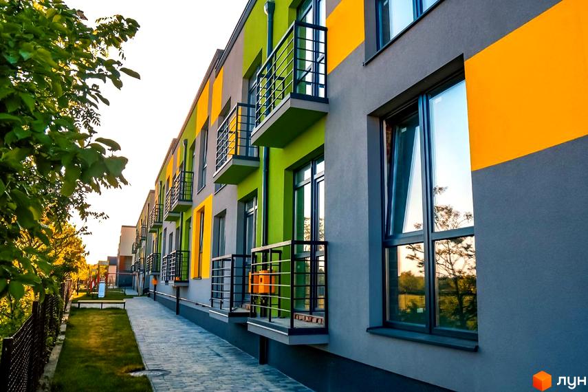 Котеджне містечко Дем’янів лаз: фасади будинків у сірих, салатових та жовтих кольорах з балконами. Прибудинкова територія має вимощену доріжку та зелені насадження.