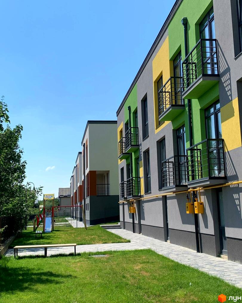 Будинки Котеджного містечка Дем’янів лаз з сірими, зеленими та жовтими фасадами, чорними балконами. Поруч доглянутий газон, дитячий майданчик та лавка.