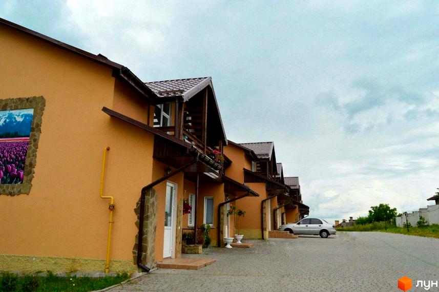 Зображення фасаду таунхаусів „Андора“ з коричневим дахом та припаркованим автомобілем. Вигляд на вулицю та зелені насадження.