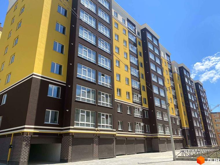 Житловий комплекс Avila Gold з жовтими та темно-коричневими фасадами, білими вікнами та цегляною основою. Перед будинком вимощена територія та ролетні ворота.
