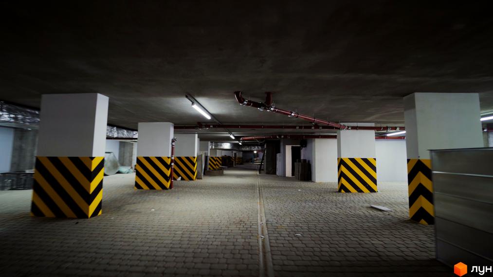 Підземний паркінг в Житловому комплексі Лісопарковий з колонами, пофарбованими в чорно-жовті смуги. На підлозі тротуарна плитка.