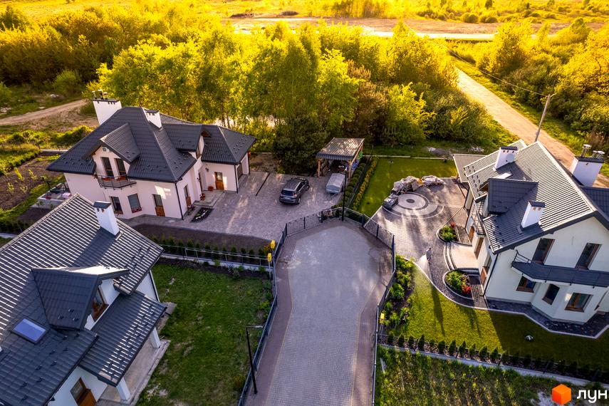 Котеджне містечко Praud Hills: елегантні будинки з темними дахами та доглянутою прибудинковою територією з газонами та бруківкою. Навколо зелені насадження та сонячне освітлення.