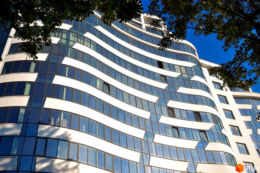 Зображення фасаду Житлового комплексу Parus з великими вікнами та білими смугами. Вигляд будівлі на фоні блакитного неба крізь зелене листя дерев.