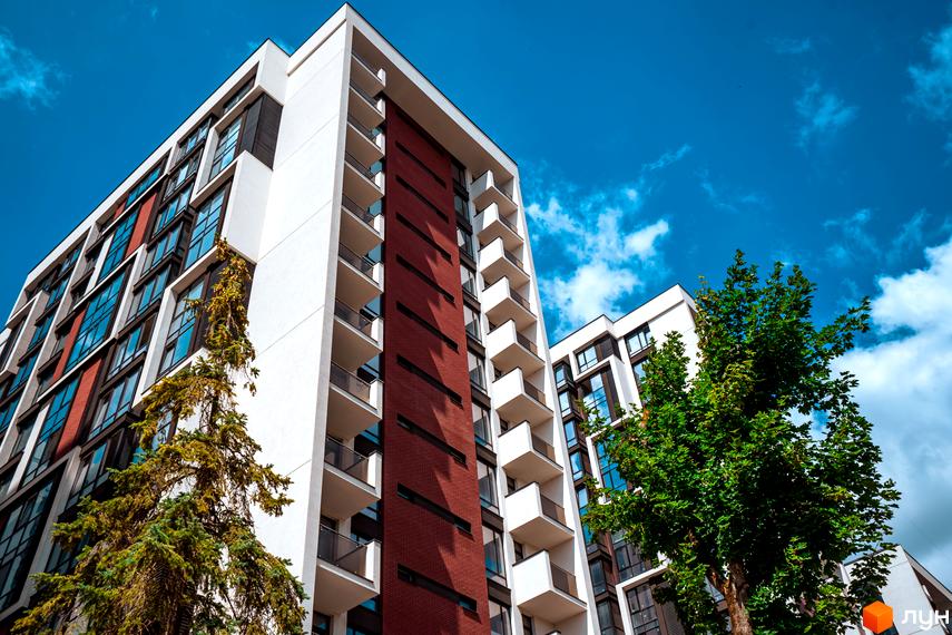 Житловий район Manhattan: сучасна архітектура з цегляними та білими фасадами, балконами та великими вікнами. Навколо будівлі зелені дерева під блакитним небом.