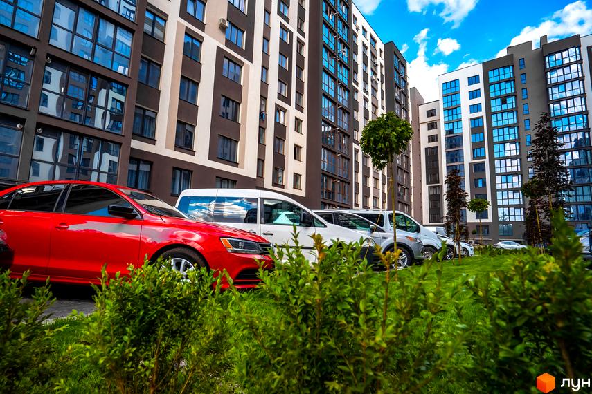 Зовнішній вигляд багатоповерхового будинку в Житловому комплексі Паркова Алея. На передньому плані прибудинкова територія з зеленими насадженнями та автомобілями.