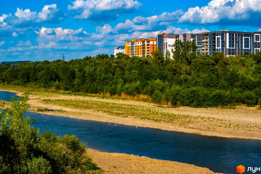 Вигляд на Житловий комплекс Паркова Алея з річки. Багатоповерхові будинки на пагорбі, навколо зелені дерева.