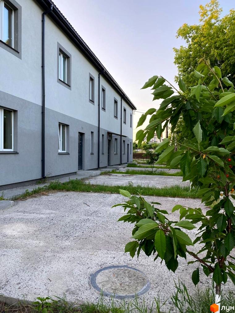 Зображення таунхаусів HarmonyBud з сірими стінами та білими вікнами. Перед будинком бетонний двір з травою та декоративне дерево.