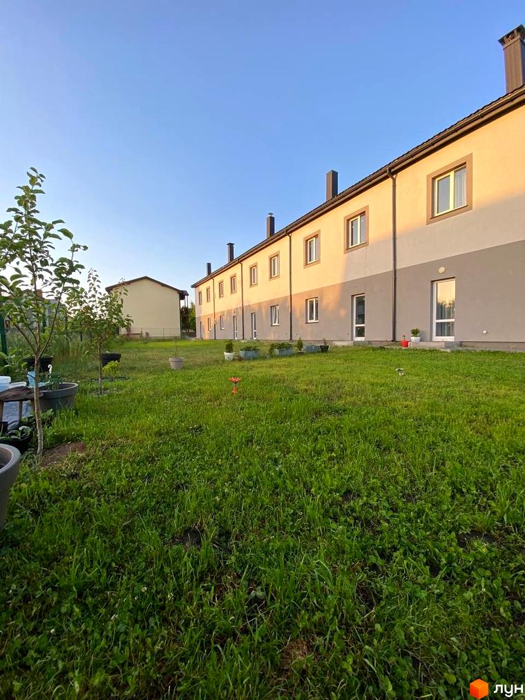 Ряд таунхаусів HarmonyBud з бежево-сірими фасадами та темними дахами. Перед будинками розкинувся зелений газон з молодими деревами та вазонами.