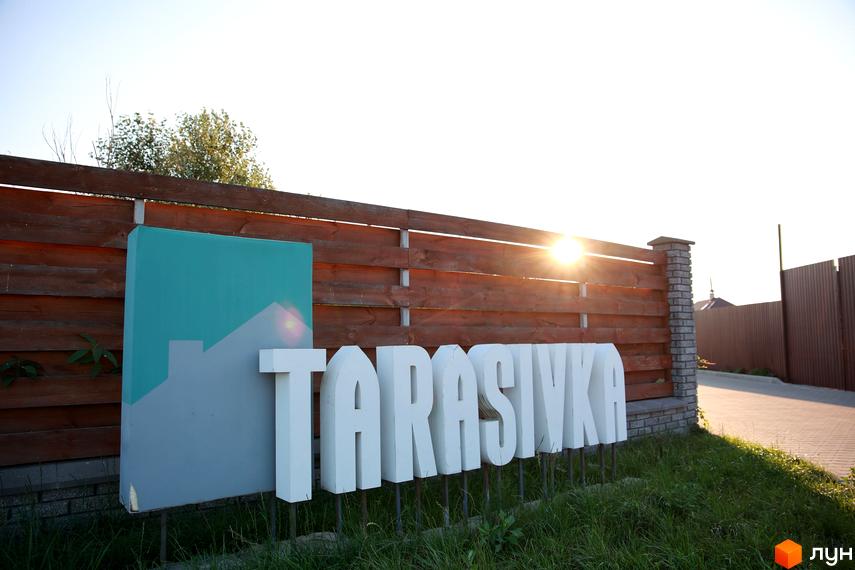 Котеджне містечко Тарасівка: фасад новобудови з великими білими літерами назви проєкту та логотипом житлового комплексу. Навколо зелена трава та дерев'яна огорожа.